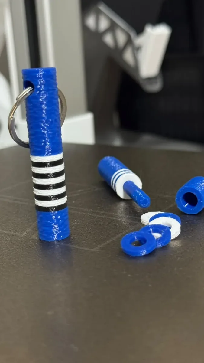 Chaveiro cilíndrico azul com anéis brancos e pretos encaixados, mostrado montado e desmontado ao lado das peças soltas.