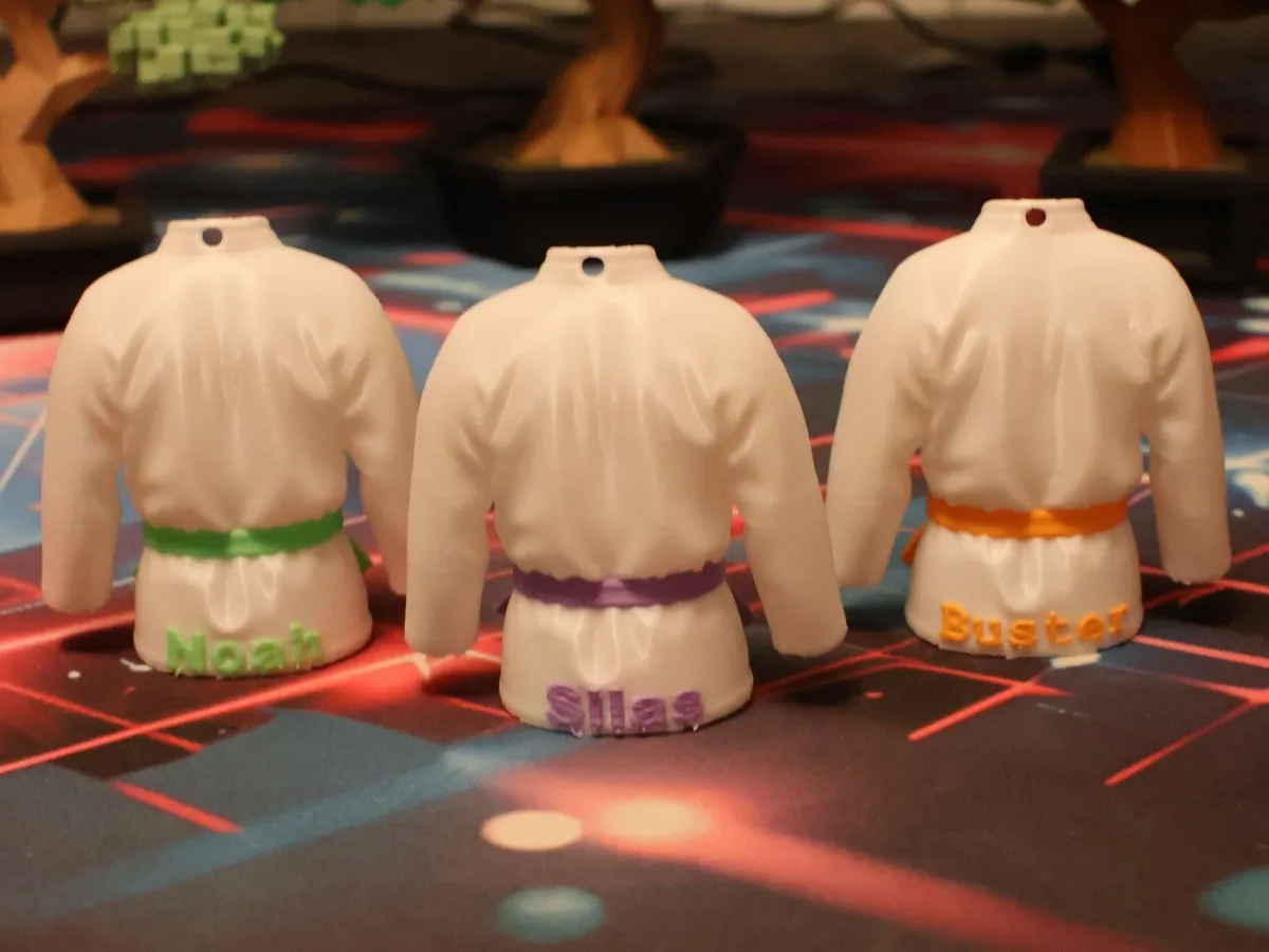 Três chaveiros em forma de kimono, vistos por trás, com os nomes Noah, Silas e Buster gravados em relevo na barra, cada um na cor da própria faixa.