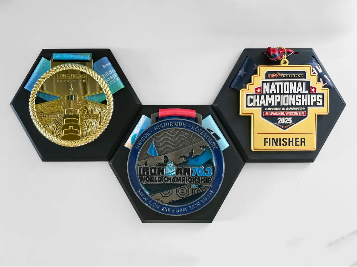 Três suportes hexagonais pretos encaixados entre si, cada um com uma medalha grande apoiada e a fita dobrada por trás.