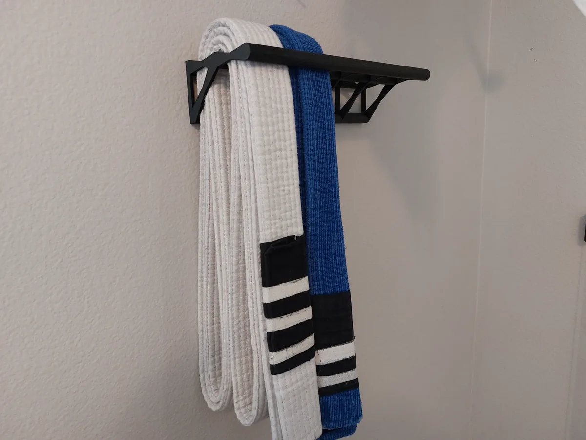 Suporte de parede preto, impresso em 3D, com estrutura vazada, segurando uma faixa branca e uma faixa azul de jiu-jitsu numa parede clara.