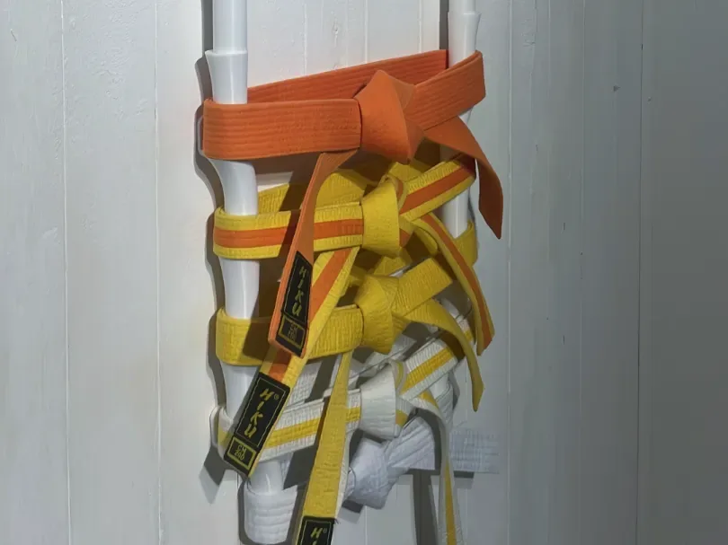 Tubo vertical branco fixado na parede, com faixas laranja, amarelas e brancas amarradas em volta dele, cada uma com o nó na frente como numa cintura.